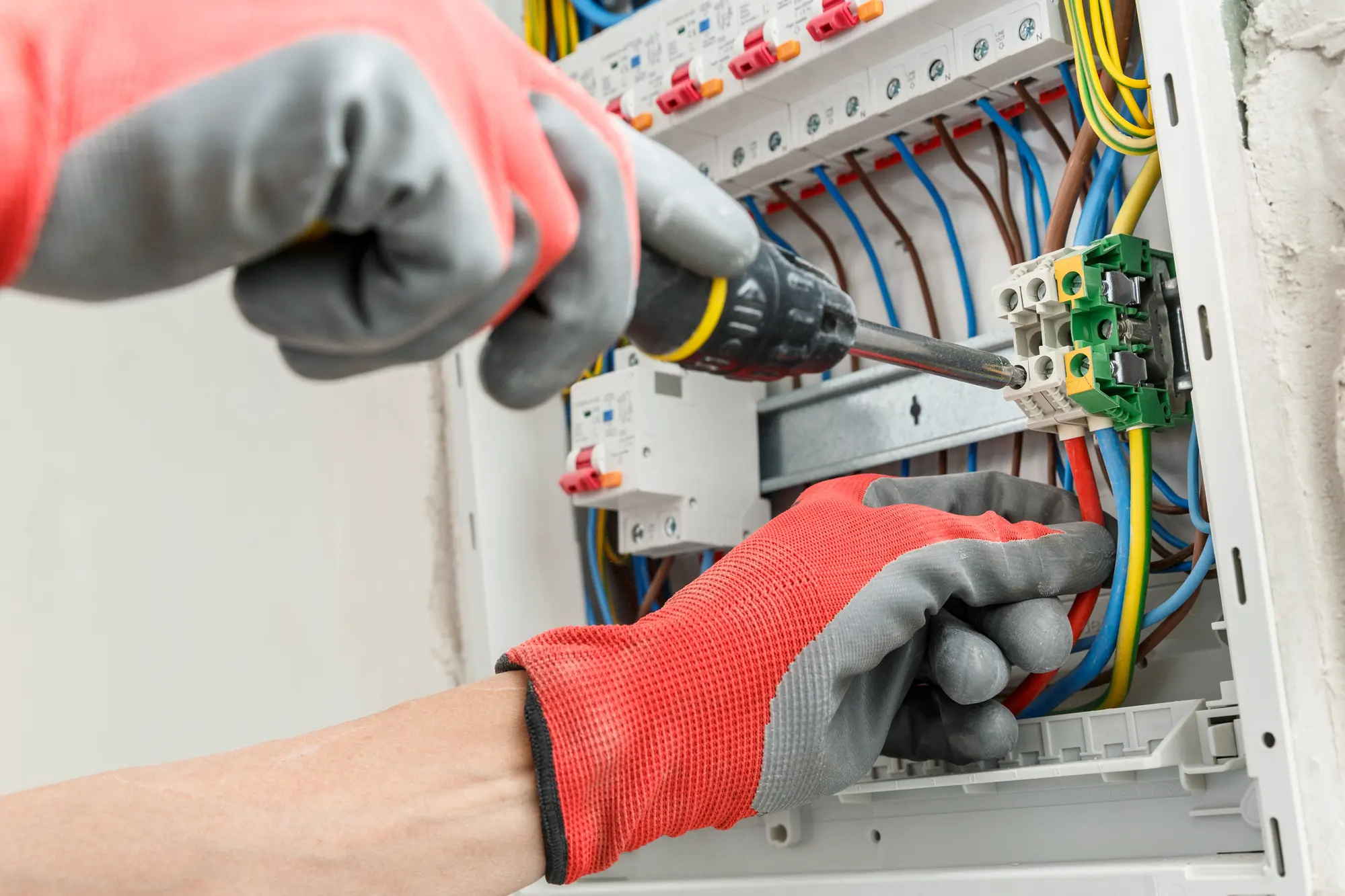 Gloved hands at an open consumer unit, one tightening a terminal with a screwdriver while the other holds the brown and blue conductors clear.