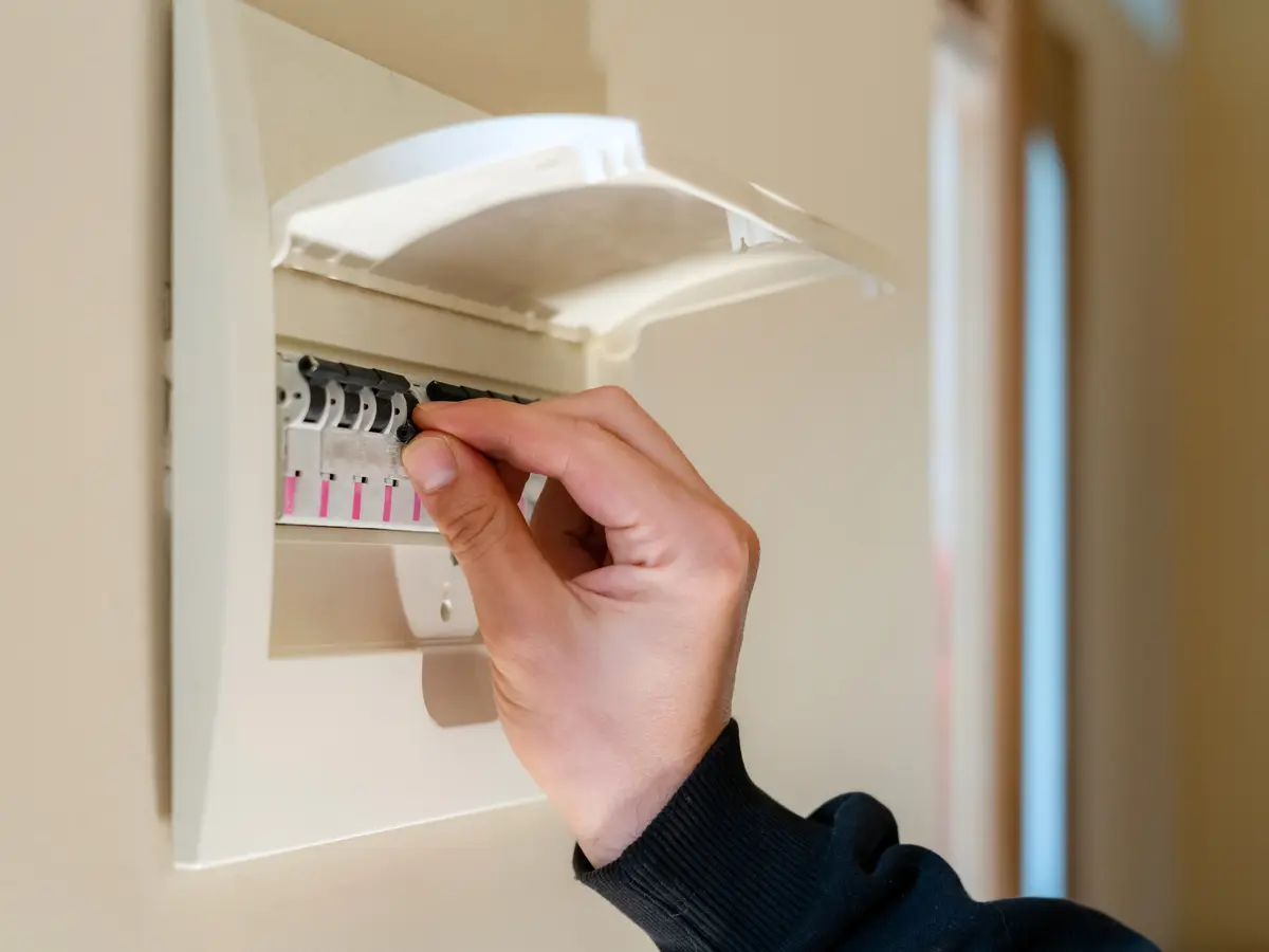 A hand resetting a breaker in a white consumer unit with its flip lid raised, on a hallway wall.