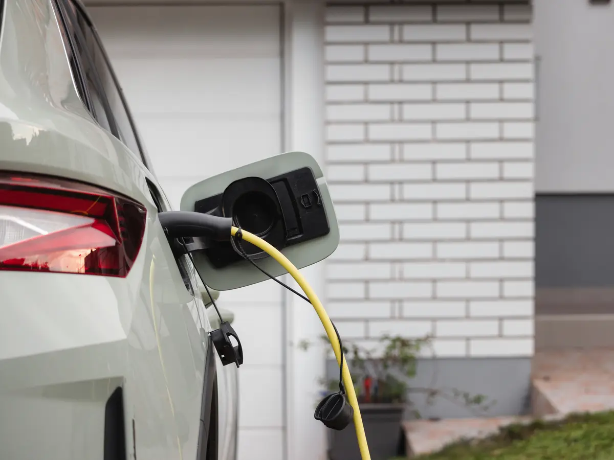 An electric car on a driveway, plugged in by a yellow charging cable beside a white brick house.