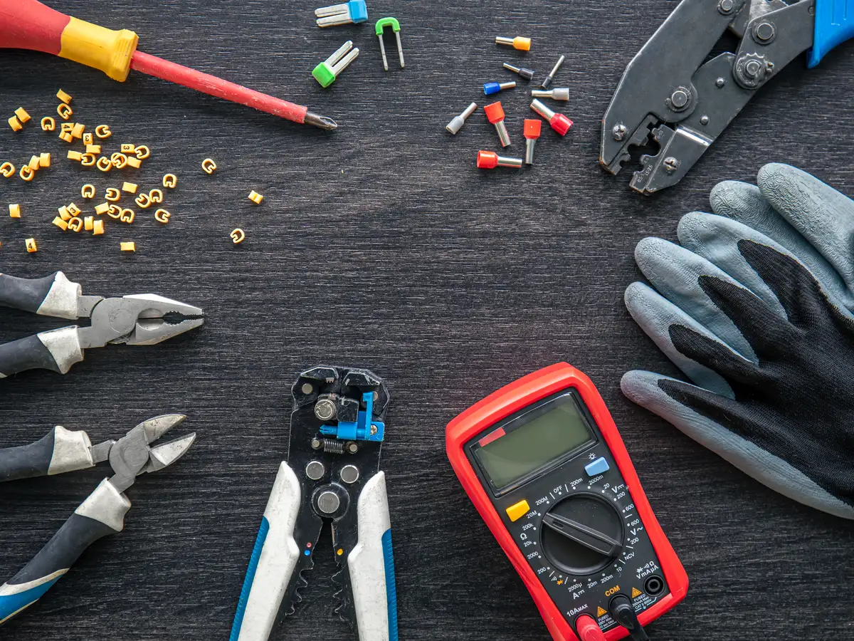 Pliers, a wire stripper, a red multimeter, gloves and crimp ferrules laid out on a dark wooden bench.