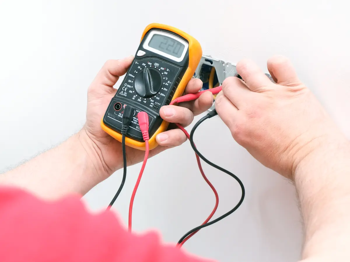 Hands holding a yellow multimeter and testing the terminals of an open wall box.
