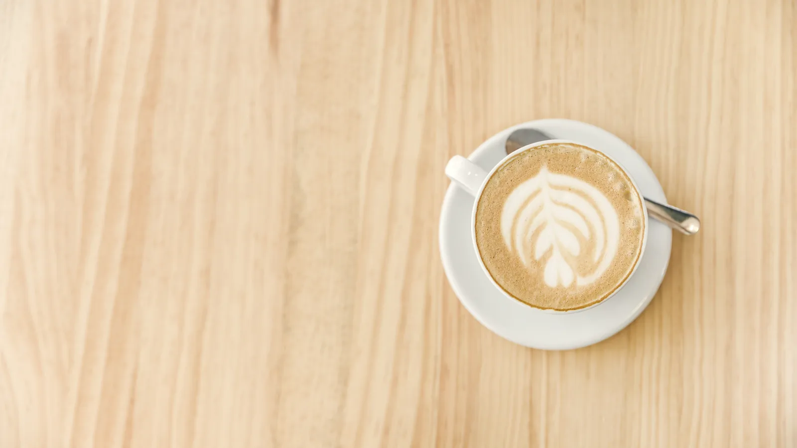 A flat white seen from above, a leaf poured in the milk, on a saucer with a spoon on a pale pine table.