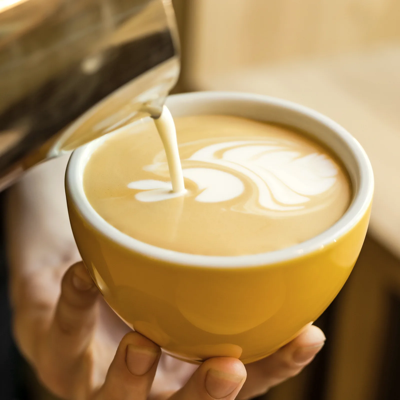 Milk poured into a yellow cup held in one hand, a rosetta forming on the crema, warm wood behind.