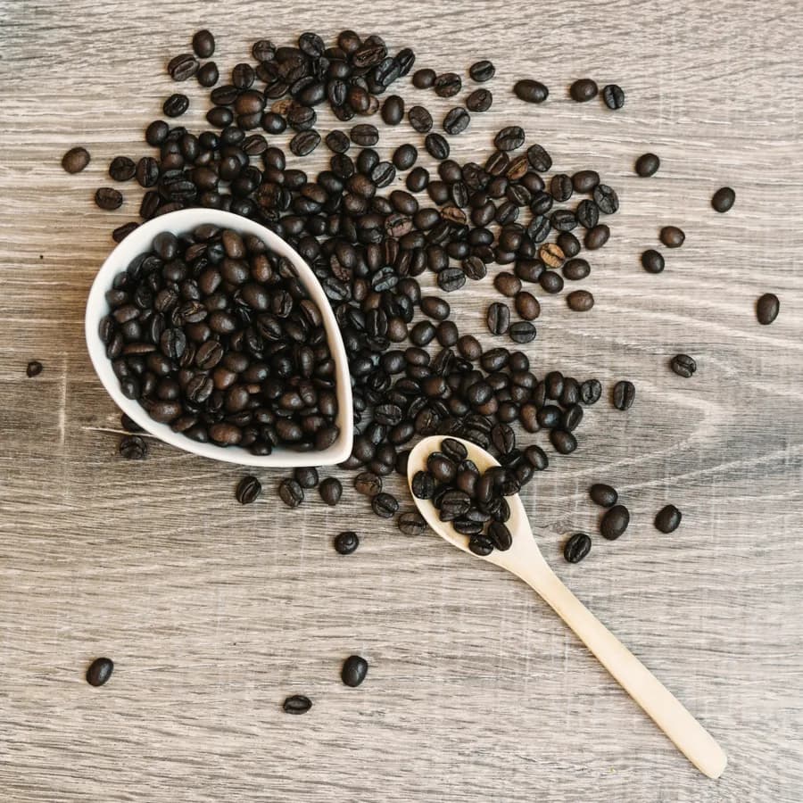 Roasted beans spilling from a white dish, with a wooden scoop, on a pale wooden table.