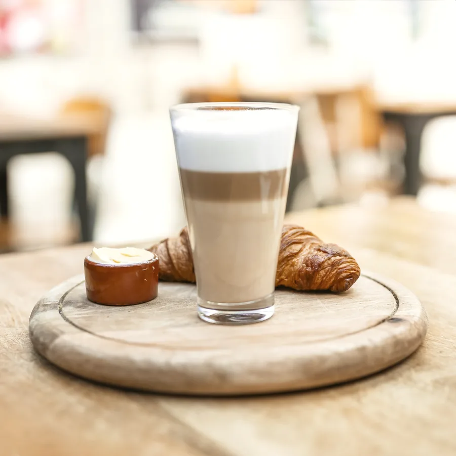 A latte macchiato in a tall glass on a wooden board, with a croissant and a small pot of butter.