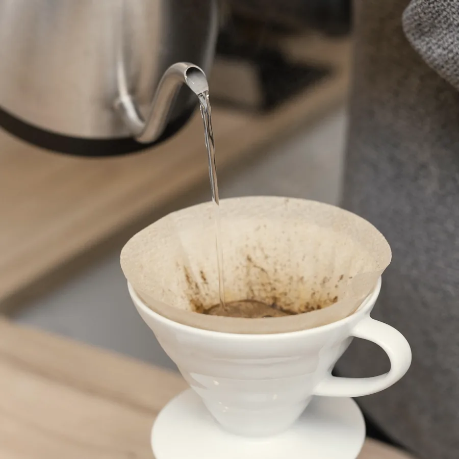 Water poured from a gooseneck kettle into a white V60 dripper lined with a paper filter.