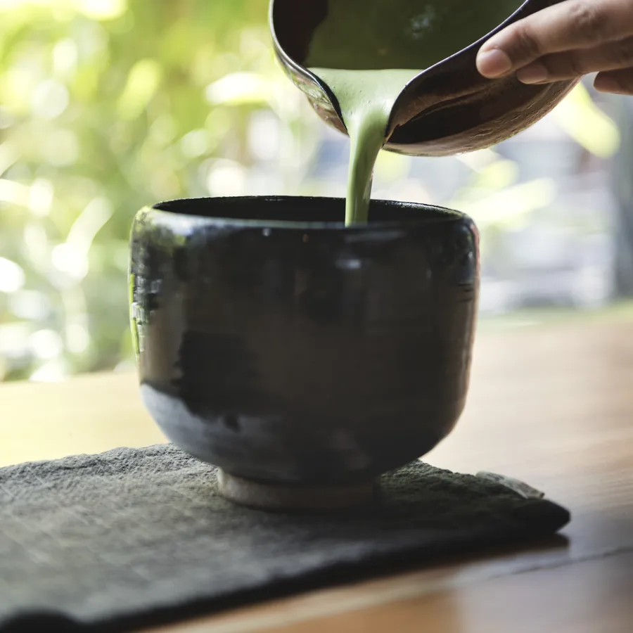 Matcha poured from a small jug into a dark stoneware bowl on a window ledge.
