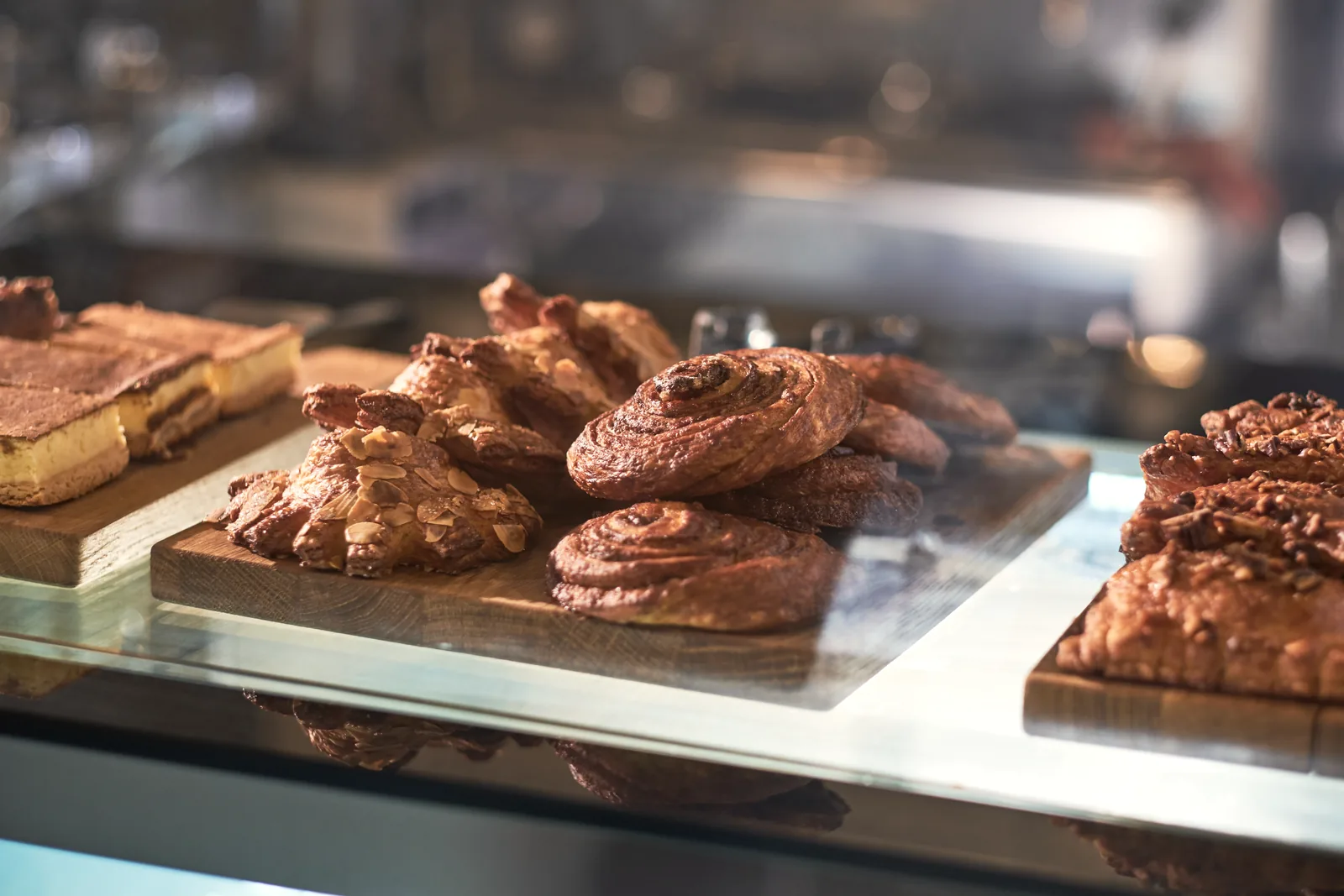 Pastries on wooden boards in a glass case: cinnamon swirls, almond croissants and custard slices in warm light.